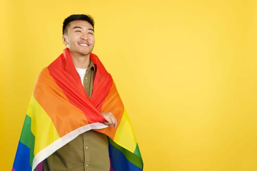 Happy chinese man wrapping with a lgbt rainbow flag in studio with yellow background