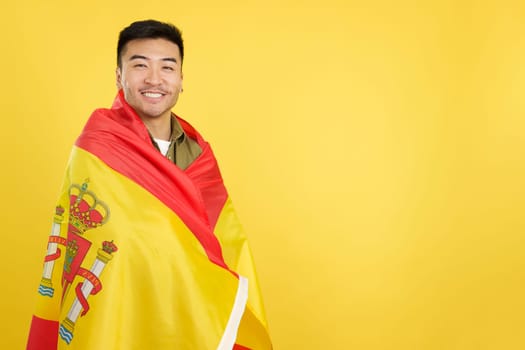 Happy chinese man looking at the camera wrapped by a spanish national flag in studio with yellow background