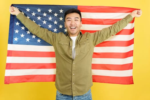 Happy chinese man raising a USA national flag while looking at camera in studio with yellow background