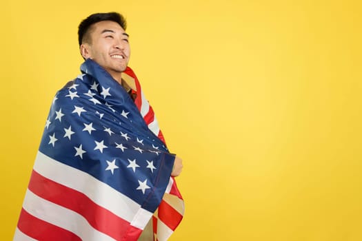 Proud chinese man wrapping with a USA national flag while looking away in studio with yellow background