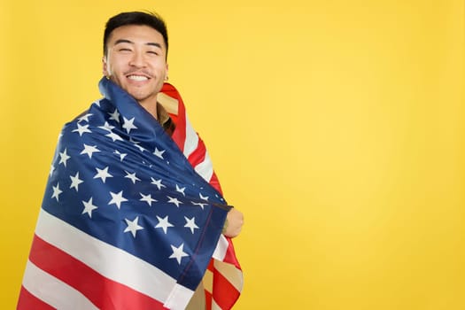 Happy chinese man looking at camera and wrapping with a United States national flag in studio with yellow background