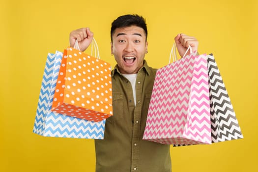 Chinese man raising many shopping bags while smiling at the camera in studio with yellow background