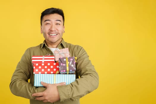 Happy chinese man holing many colorful gifts in studio with yellow background