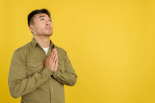 Chinese man praying looking up with hands folded in studio with yellow background