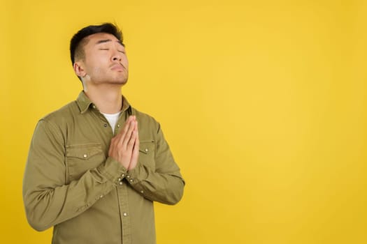 Chinese man praying with hands folded and eyes closed in studio with yellow background
