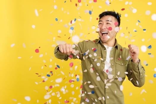 Happy chinese man surrounded by confetti flying in the air in studio with yellow background