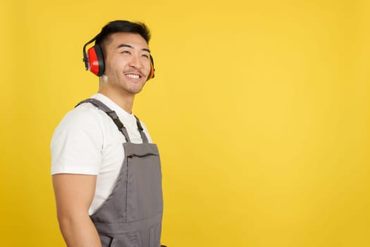 Chinese carpenter smiling distracted while looking up and wearing protective gear and headphones in studio with yellow background