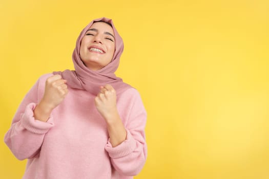 Happy muslim woman celebrating while raising fists in studio with yellow background