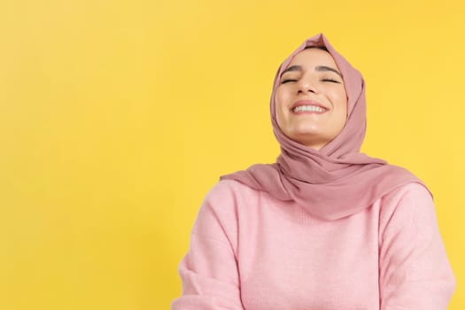 Happy muslim woman laughing with eyes closed in studio with yellow background