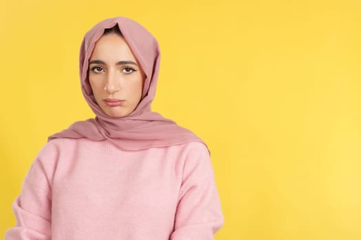 Sad muslim woman wearing head scarf looking at camera in studio with yellow background