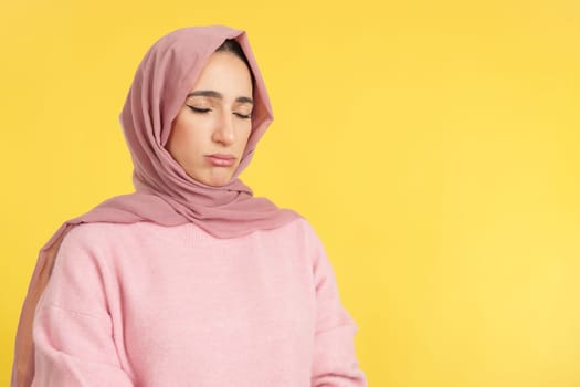 Sad muslim woman standing with eyes closed in studio with yellow background