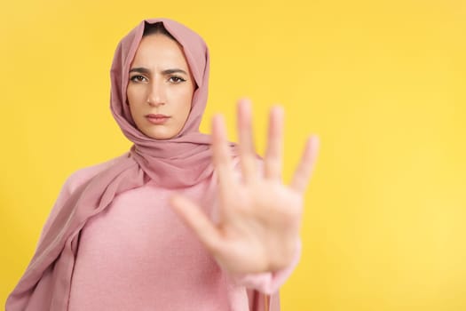 Muslim woman gesturing prohibition with hand in studio with yellow background