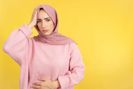 Muslim woman gesturing bodily discomfort and headache in studio with yellow background