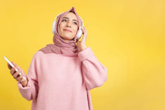 Happy muslim woman smiling while listening to music with the mobile and headphones in studio with yellow background