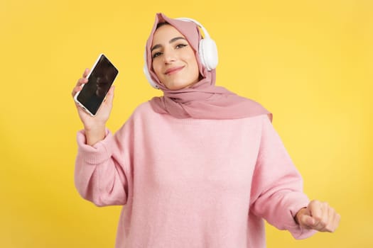 Happy muslim woman dancing and listening to music with mobile in studio with yellow background