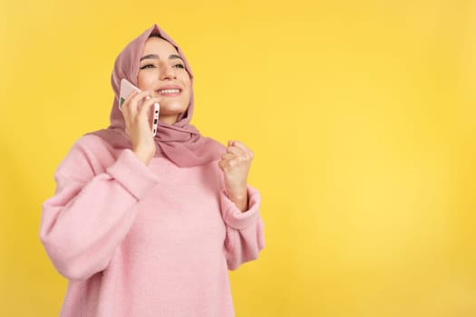 Happy muslim woman celebrating while talking to the mobile phone in studio with yellow background