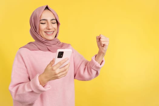 Excited muslim woman celebrating raising the fist while using a mobile in studio with yellow background