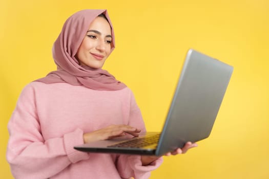Happy muslim woman standing using a laptop in studio with yellow background