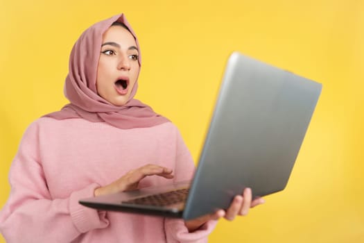 Surprised muslim woman with head scarf using a laptop in studio with yellow background