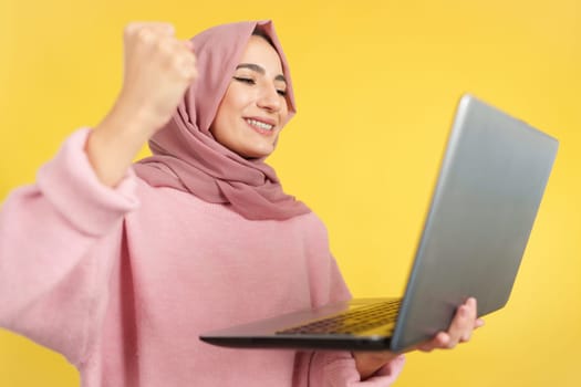 Happy muslim woman celebrating while using a laptop in studio with yellow background