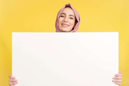 Happy muslim woman with scarf holding a blank panel in studio with yellow background