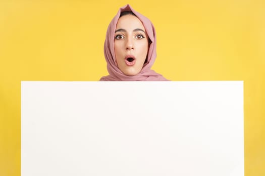 Surprised muslim woman with scarf holding a blank panel in studio with yellow background
