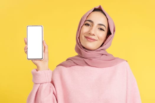 Happy muslim woman holding a mobile with a blank screen in studio with yellow background