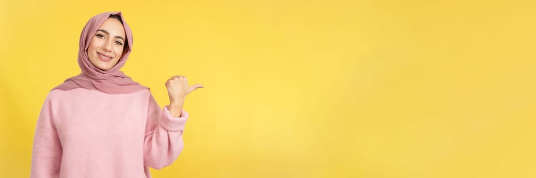 Muslim woman smiling at camera and pointing to the side in studio with yellow background