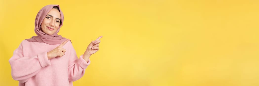 Muslim woman indicating with finger the blank space to the side in studio with yellow background