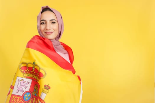 Happy muslim woman looking at camera while wrapping with spanish national flag in studio with yellow background