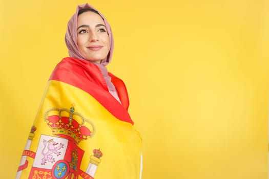 Dreamy muslim woman wrapped by a spanish national flag in studio with yellow background