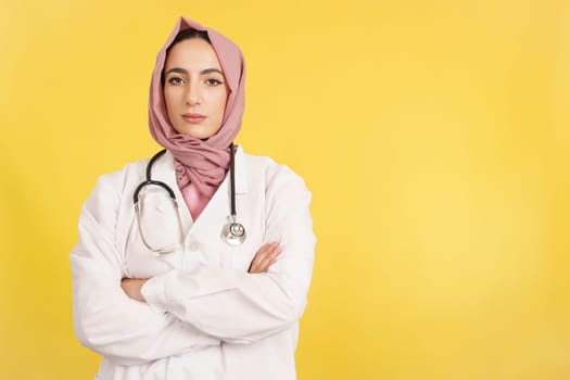 Serious female muslim doctor with arms crossed looking at camera in studio with yellow background