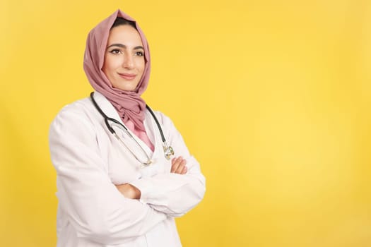 Happy female muslim doctor with arms crossed looking away in studio with yellow background