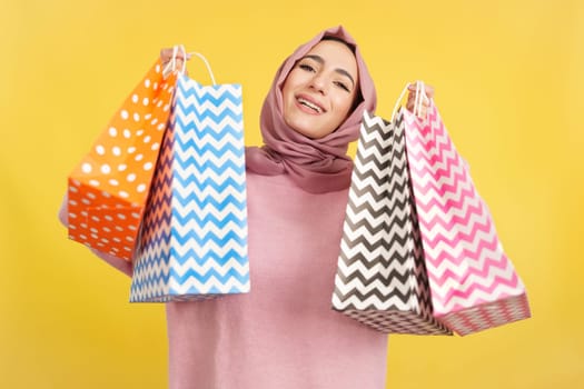 Muslim woman smiling at the camera while showing shopping bags in studio with yellow background