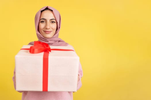 Happy muslim woman smiling at camera while giving a gift in studio with yellow background