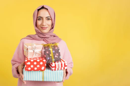 Muslim woman smiling with many gifts in the hands in studio with yellow background