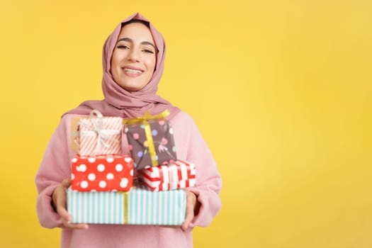 Happy muslim woman with scarf holding many gifts in studio with yellow background