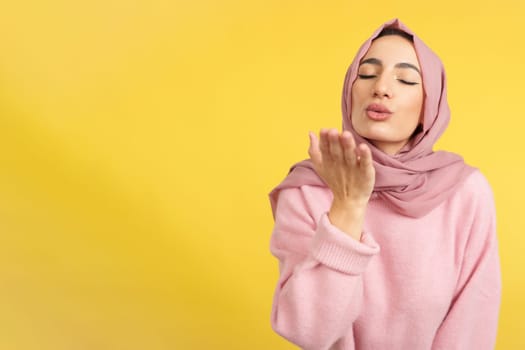 Muslim woman blowing a kiss with eyes closed in studio with yellow background