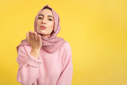 Muslim woman blowing a kiss while looking at the camera in studio with yellow background