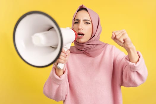 Angry muslim woman screaming using a loudspeaker in studio with yellow background