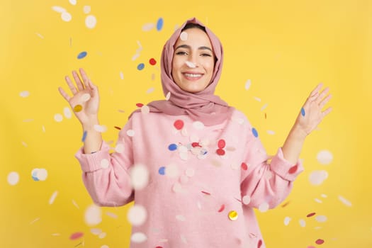 Happy Muslim woman surrounded by confetti flying in the air in studio with yellow background