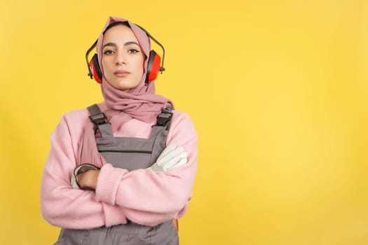 Serious muslim female carpenter in work uniform looking at camera in studio with yellow background