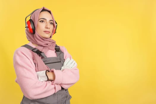 Serious Muslim female carpentry worker standing with arms crossed with work uniform in studio with yellow background