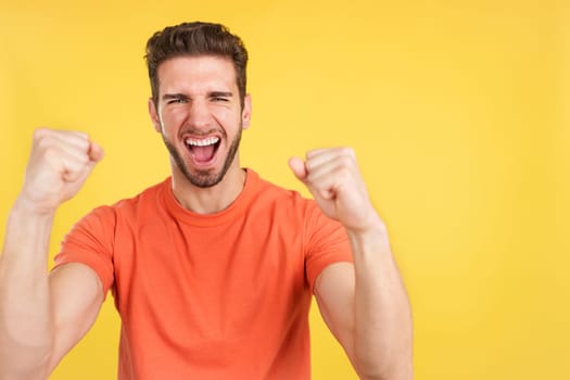 Happy caucasian man celebrating while raising fists in studio with yellow background