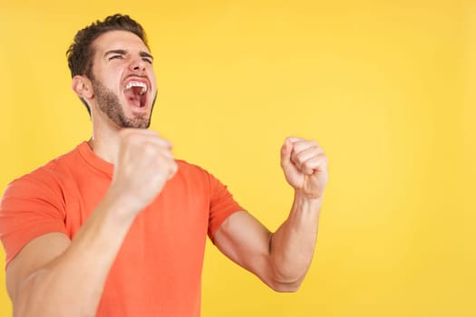 Excited caucasian man celebrating while raising arms in studio with yellow background