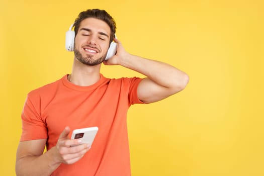 Caucasian man listening to music with the eyes closed using a mobile phone and headphones in studio with yellow background