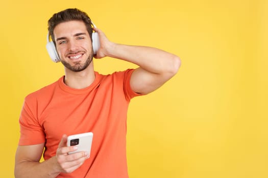 Caucasian man listening to music with headphones and a mobile in studio with yellow background