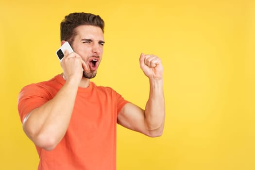 Happy caucasian man celebrating while taking to the mobile in studio with yellow background