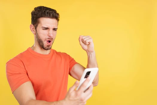 Excited caucasian man celebrating while using the mobile phone in studio with yellow background