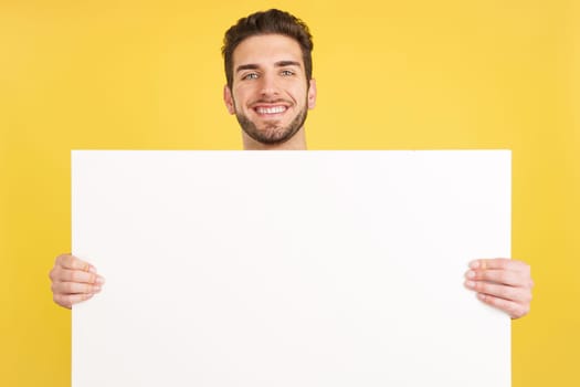 Happy caucasian man smiling while holding a blank panel in studio with yellow background